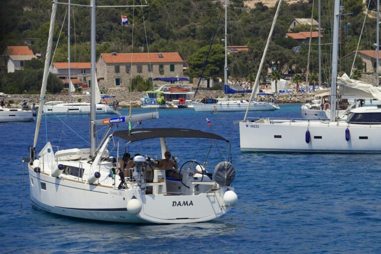 Sailing yachts in a beautiful Mediterranean bay with houses and lush greenery in the background.