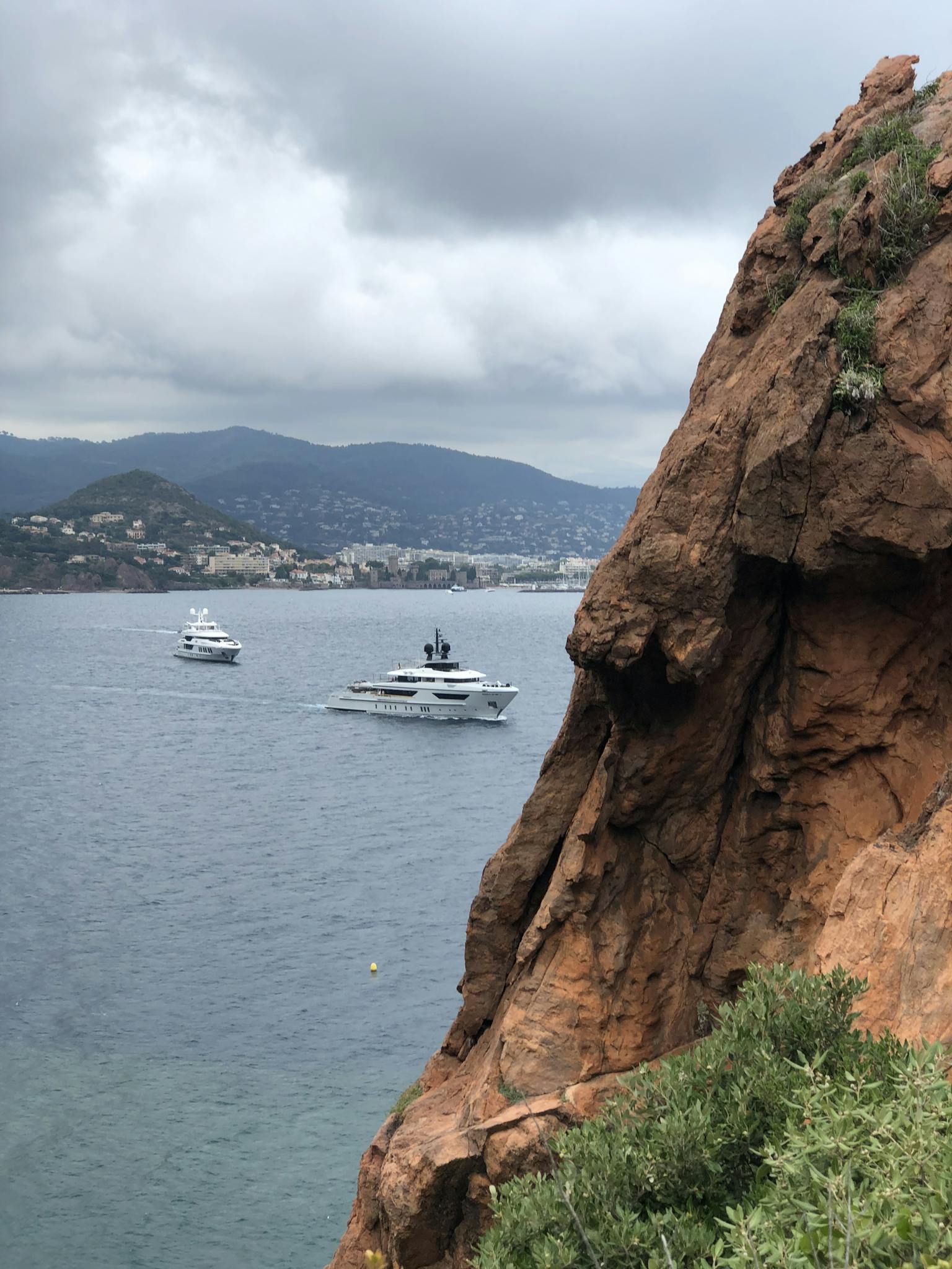 Scenic view of luxury yachts sailing on the Mediterranean Sea near rugged cliffs under cloudy skies.